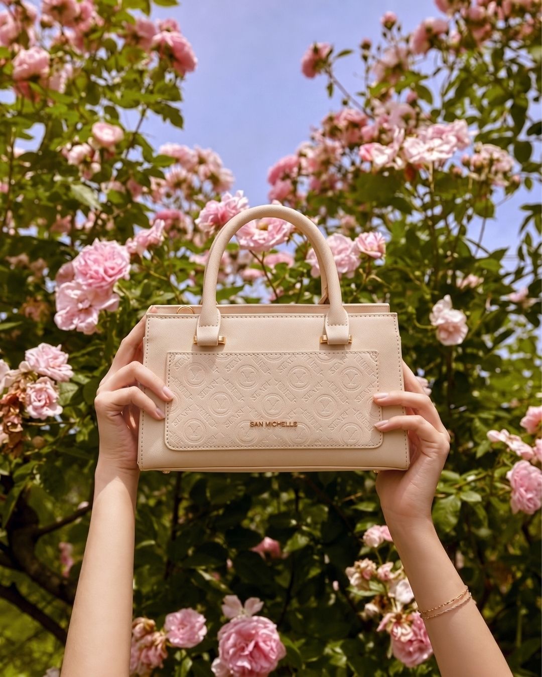 Hands holding up a blush pink San Michelle handbag among pink roses