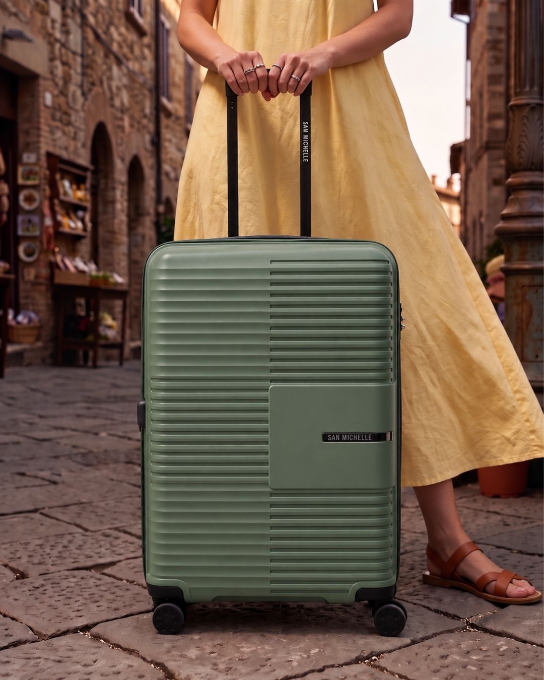 Woman in a yellow dress with a sage green hard shell checked suitcase on a cobbled street